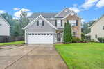 A two-story suburban house at 2134 Westbourne Drive, Creedmoor, features gray siding, brick accents, a white double garage door, and a well-kept lawn, pictured on a cloudy day with a wet driveway.
