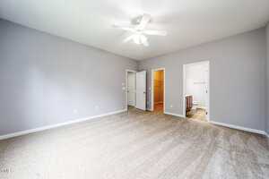 A spacious, empty bedroom with light gray walls, beige carpet, a white ceiling fan, and two open doorways leading to a walk-in closet and a bathroom. A closed white door is also visible on one wall.
