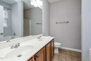A bathroom with a double-sink vanity, large mirror, wooden cabinets, a toilet, towel bar, and a bathtub with a shower curtain, featuring gray walls and tile flooring.