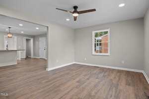 A spacious, empty room with light beige walls, wood-style flooring, a ceiling fan with a light, recessed ceiling lights, and a window showing part of a brick building, with a view into a kitchen area.