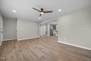 A spacious, empty room with wood flooring, beige walls, a ceiling fan with light, recessed lighting, and an open doorway leading to a modern kitchen area with white cabinetry.