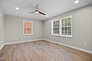 Empty room with light gray walls, wood-style flooring, a ceiling fan with light, recessed ceiling lights, and two windows letting in natural light. An additional window faces a brick exterior wall.