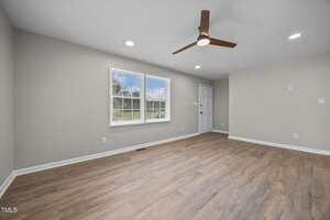 Empty room with light gray walls, wood-style flooring, a ceiling fan with three blades, recessed lighting, a large window, and a white door in the background.