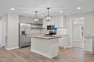 A modern kitchen with white cabinets, stainless steel appliances, granite countertops, a wood-topped island with a sink, pendant lights above the island, and light wood flooring.