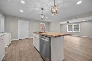 A modern kitchen with light wood flooring, a central island with a wooden countertop and sink, stainless steel dishwasher, white cabinets, pendant lights, and an open layout leading to a bright living area.