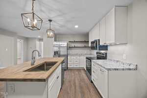 A modern kitchen with white cabinets, a wooden island with a sink, stainless steel appliances, marble countertops, open shelving, and pendant lights hanging from the ceiling. The floor is a light wood finish.