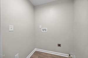 An empty laundry area with beige walls, white baseboards, wood-look flooring, water hookups, an electrical outlet, and a dryer vent on the right side of the back wall.