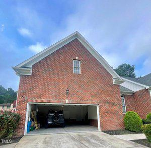 A brick house with a double garage door open; a vehicle is parked inside the left bay, and the right bay is empty. Lawn equipment and shelves are along the garage walls. Bushes and a driveway are visible in front.