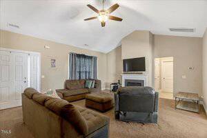 Living room with brown sofas, a matching ottoman, and a black recliner. A TV is mounted above a white fireplace. The walls are beige, carpet is brown, and a ceiling fan with light hangs from the vaulted ceiling.