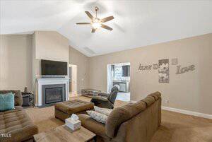 A living room with brown carpet, two brown sofas, a coffee table, a wall-mounted TV above a white fireplace, ceiling fan, and wall art reading "home" and "love." The kitchen is visible through a doorway.