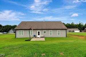 Single-story gray house with a gable roof, several windows, a small concrete patio with a grill and chair, and a basketball hoop in the backyard, set on a grassy lawn with neighboring houses visible.