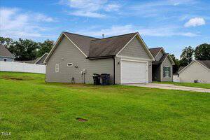 A single-story house with gray siding, attached two-car garage, paved driveway, and trash bins beside the garage, set on a green lawn under a partly cloudy sky. Fence and neighboring houses are visible in the background.