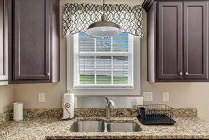 A kitchen sink with a granite countertop, paper towel holder, soap dispenser, drying rack, and dark wood cabinets. A window with blinds and a patterned valance is above the sink, letting in natural light.