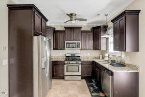 Modern kitchen with dark wood cabinets, stainless steel appliances including a refrigerator, oven, microwave, and dishwasher, granite countertops, beige tile floor, ceiling fan, and window with a patterned valance.