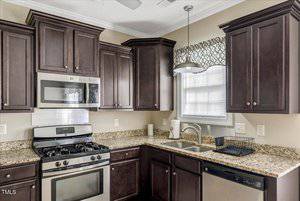 A kitchen with dark wood cabinets, granite countertops, a stainless steel gas stove with oven, microwave, dishwasher, double sink, toaster, and a window with a patterned valance above the sink.
