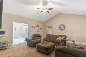 A living room with beige walls, brown carpet, and a ceiling fan. There are two armchairs, a sofa, and an ottoman. Decorative wall art and a clock hang on the wall. An open doorway leads to another room.