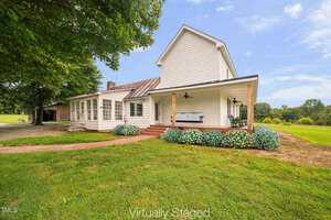 A white two-story farmhouse with a front porch, ceiling fans, and hydrangea bushes, set on a grassy lawn with trees and an open field in the background. The image is labeled "Virtually Staged.