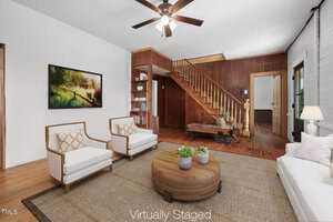 A living room with a wooden staircase, white walls, wood-paneled accent wall, and ceiling fan. Virtually staged with white armchairs, a white sofa, a round wooden coffee table, and wall art. A rug covers the floor.