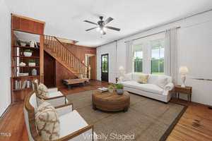 A virtually staged living room with wood floors, a white sofa, two white armchairs, a round coffee table, and built-in shelves. Wooden stairs lead to an upper level, and large windows let in natural light.