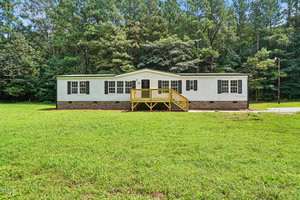 A single-story manufactured home at 330 Sycamore Lane in Henderson features beige siding, black shutters, a small yellow front porch with steps, a spacious grassy yard, and dense trees in the background.