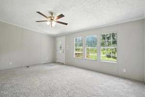 A carpeted living room with light gray walls, a white ceiling fan with lights, three large windows showing green trees outside, and a white front door with an oval window.