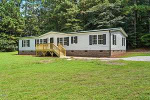 A single-story house with white siding, black shutters, and a brick foundation is set on a grassy lot with a small porch and surrounded by trees. There is a gravel area near the house.