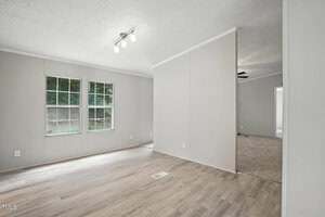 A bright, empty room with light-colored wood flooring, two large windows on the left wall, white walls, and a ceiling light fixture. An open doorway leads to a carpeted area in the background.