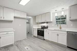 A modern kitchen with white cabinets, light gray walls, light wood flooring, stainless steel appliances, a window above the sink, and recessed lighting. An open doorway leads to a pantry or closet.