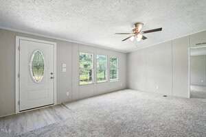 A bright, empty room features light gray walls, a white front door with an oval window, three large windows, a ceiling fan with lights, and carpeted flooring. A section of the floor near the door is vinyl.