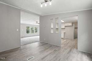 A view of a modern, unfurnished open-plan home interior with light gray walls, wood-look flooring, and white trim. The space includes a living area, dining area, and a kitchen with white cabinets. Large windows provide natural light.