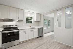 A modern kitchen with white cabinets, stainless steel oven and dishwasher, light countertops, two pendant lights, and windows overlooking greenery. Light wood flooring extends into an adjacent room.