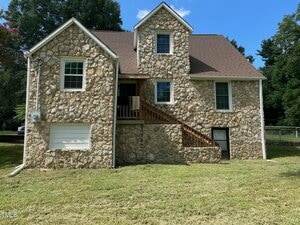 A two-story stone house with a brown roof, multiple windows, a small upper balcony with wooden railing, a white garage door, and a grassy yard. Trees are visible in the background under a blue sky.