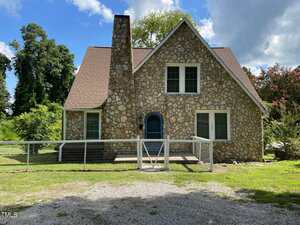 A stone house at 1203 Dorsey Avenue in Henderson features a steep roof, two chimneys, arched front door, and metal fence. Several windows overlook grass, trees, and shrubs under a partly cloudy sky.