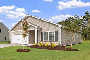 Single-story house with beige siding, white trim, and stone accent under front windows. Attached two-car garage on the left. Small landscaped front yard with shrubs and two young trees. Blue sky with scattered clouds.