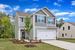Two-story gray house with white trim, double garage door, small covered front porch, and landscaped yard with a young tree. Blue sky with scattered clouds in the background.