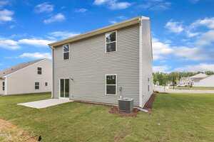 A two-story house with light gray siding, several windows, a concrete patio, and a central air conditioning unit in a newly landscaped yard on a clear day. Neighboring houses are visible in the background.