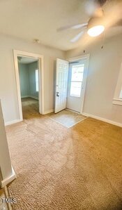 A small room with beige carpet, white walls, a ceiling fan with a light, an exterior door with a window, and an open doorway leading to another room. Sunlight filters through the window.