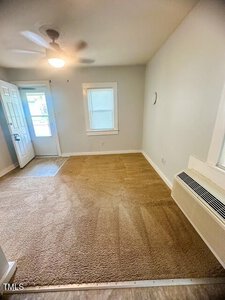 A small, unfurnished room with beige carpet, light gray walls, a window, a door with glass panels, a ceiling fan with a light, and a wall-mounted air conditioning unit. Natural light is coming through the door.