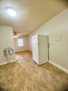 A white refrigerator stands against a wall in a kitchen with beige flooring and light-colored walls. An open area with a window, ceiling fan, and a dining space is visible in the background.