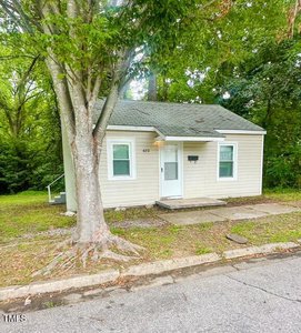 A small beige house with a green roof and a white front door sits close to the street, shaded by a large tree, with a sidewalk and grass yard in front and dense trees in the background.