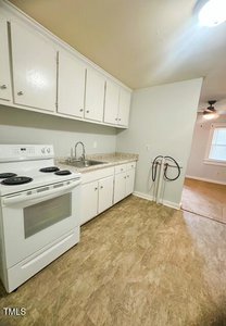 A small kitchen with white cabinets, a white electric stove, a countertop with a single sink, and beige flooring. An open doorway leads to a carpeted living area with a ceiling fan and window.