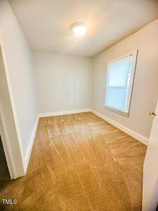 A small empty room with beige carpet, white walls, a single window with blinds, and a ceiling light fixture. The room appears clean and well-lit.