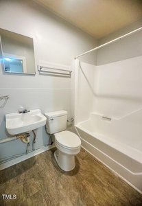 Small bathroom with a white sink, toilet, and bathtub-shower combo. A mirror hangs above the sink, with an empty towel rack on the wall. The floor is covered with tan vinyl flooring.