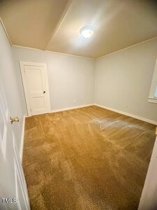 Empty room with beige carpet, white walls, a closed white door, a ceiling light, and a window on the right wall. Room appears clean and well-lit.