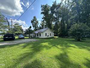 A small single-story white house with a front porch sits on a grassy lawn surrounded by trees. Two vehicles are parked on a gravel driveway to the left, under a sunny blue sky with scattered clouds.