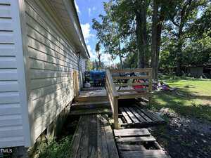A wooden ramp and steps lead to a small deck attached to the side of a light-colored house. The yard is grassy with scattered items, and trees provide shade in the background under a blue sky.
