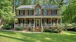 A two-story brick house at 4137 Grey Fox Court, Oxford, features a prominent central window, covered front porch with white railings, and steps to the lawn. Mature trees and shrubs surround the home, with flower pots lining the porch and walkway.