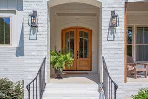 A wooden double front door with glass panels is centered on a white brick porch. Two wall-mounted lanterns flank the door, and a potted fern sits to the left. Black metal railings line the steps leading up to the entry.