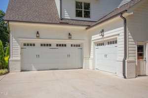 Two white garage doors and a side entry door on a modern house exterior with white siding, brick trim, outdoor lights, and a clean concrete driveway.