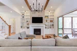 A living room with a beige sectional sofa facing a fireplace with a mounted TV, built-in shelves on both sides, wood ceiling beams, a large black chandelier, and sliding glass doors leading to a screened porch.